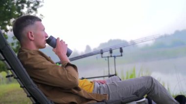 Genç, şık balıkçı bir adam balıkçı koltuğunda oturup bir içki içiyor ve termostan çay içiyor. Göl oltalarının ve ağaçların arka planında doğa var. Hipster adam tatilde. Etkin yeniden yaratma 