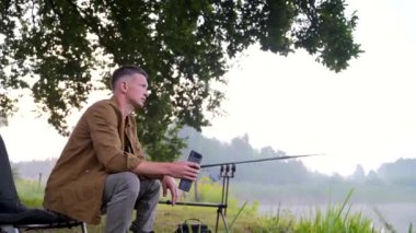 Genç, şık balıkçı bir adam balıkçı koltuğunda oturup bir içki içiyor ve termostan çay içiyor. Göl oltalarının ve ağaçların arka planında doğa var. Hipster adam tatilde. Etkin yeniden yaratma 