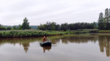 Genç bir adam şişme bir teknede oturur ve göl ya da nehrin ortasındaki suya bir olta atar. sazlıkların ve ormanın arka planı. Hipster adam balıkçı aktif eğlence, kamp