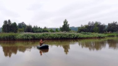 Genç bir adam şişme bir teknede oturur ve göl ya da nehrin ortasındaki suya bir olta atar. sazlıkların ve ormanın arka planı. Hipster adam balıkçı aktif eğlence, kamp