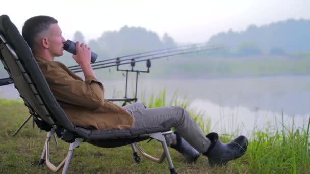 Young stylish fisherman guy sitting on a fishing chair and drinking a drink, tea coffee from a thermos. background of lake fishing rods and trees nature. Hipster man on vacation. Active recreation — Stock Footage Young Stylish Fisherman Guy Sitting Fishing Chair Drinking Drink Tea Video Clip