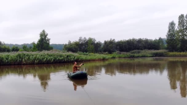 年轻人坐在充气船上 把纺纱或钓竿扔进湖中或河里的水里 芦苇和森林的背景 嬉皮士人渔夫积极娱乐露营 — 图库视频影像