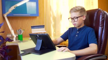 Genç ve çekici bir öğrenci masada tabletle online ders veriyor. Evde uzaktan öğrenme. Odada gözlüklü bir genç internette bir cihaz kullanıyor.