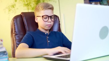 Çevrimiçi takılırken dizüstü bilgisayarla ödev yapan, mavi tişört giyen ve evde gözlüklü genç bir çocuk. Öğrenmek ve bilgi aramak için bilgisayarı kullanan genç.