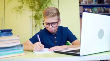Küçük tatlı bir çocuğun portresi bir dizüstü bilgisayarla masada oturmuş ders veriyor ve bir defter görevi yazıyor. Gözlüklü genç evde uzaktan öğrenme üzerine ev ödevi yapıyor.
