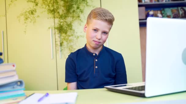 疲れた若い金髪の少年の肖像画は 自宅のテーブルでノートパソコンとノートパソコンでレッスンを教えています 十代の少年は勉強中に碑文の助けを借りて画像を示しています 顔の近くに疲れ — ストック動画