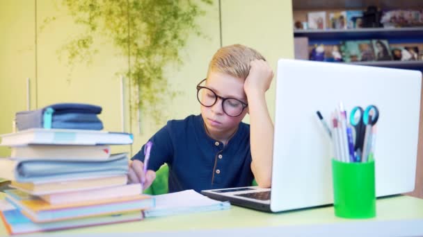 可爱疲惫的少年男孩坐在桌旁 在家中远程学习时 带着眼镜 在笔记本上写字 在网上读书和买白色笔记本电脑的过程中 无聊的孩子和学生 — 图库视频影像