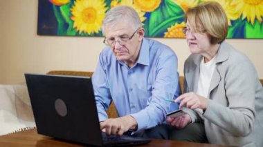 Oturma odasında oturan sevimli yaşlı çift dizüstü bilgisayarında kredi kartıyla online ödeme yapıyor. Büyükanne ve büyükbaba internetten alışveriş yapıyor..