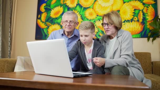 可爱的金发男孩和祖父母坐在沙发上 在五颜六色的客厅里用白色笔记本电脑展示一些东西 青少年花时间与老一辈人在一起 使用孤立的计算机 — 图库视频影像