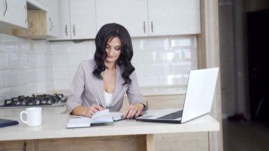 Esmer iş kadını mutfakta karantina sırasında mesafe çalışması yapıyor. Makyajlı ve günlük kıyafetleri olan çekici bir kız. Not alıyor. Sonra da evde dizüstü bilgisayarda yazıyor..