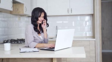 Güzel, gülümseyen bir kadın, parlak beyaz bir mutfakta dizüstü bilgisayarla yazarken akıllı telefonuyla konuşuyor. Esmer, makyajlı, bilgisayarla çalışan ve telefonla konuşan..