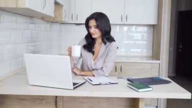 Genç çekici esmer iş kadını mutfakta oturuyor, kahve içerken laptop ve belgelerle çalışıyor. Bilgisayarda daktilo eden kadın, karantina sırasında izole edilmiş not defterine bir şey yazıyor.