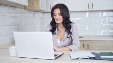 Genç ve çekici esmer iş kadını evinde, şık bir mutfakta dizüstü bilgisayarla video konferansı yapıyor. Başarılı, kendine güvenen bir kadın çalışıyor ve kapalı alanda web kamerasıyla ağ yapıyor..