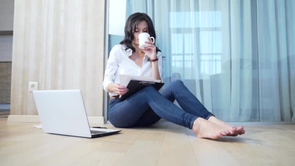 身穿休闲装的年轻女商人坐在房间的地板上 用手提电脑电话和文件工作 在家远离在线工作 Brunette女商人在互联网上做自由职业 — 图库视频影像