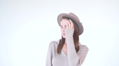 Şapkalı, saçlarına dokunan, gülümseyen ve beyaz arka plandaki kameraya bakan genç, sevimli bir kız. Stüdyoda poz veren çekingen, güzel, kahverengi saçlı bir kadının yakın portresi..