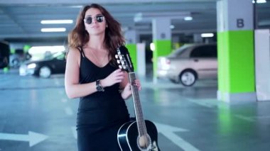 ... siyah şık elbiseli ve güneş gözlüklü, modaya uygun, genç bir bayan gitarist, yürüyen ve otoparkta kameraya bakan genç ve şık bir bayan. Şehir arkaplanı