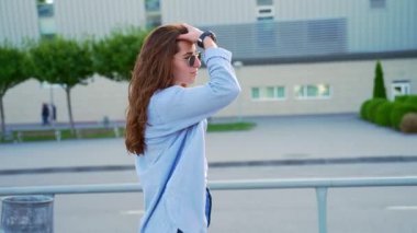 Güneş Gözlüklü Kıvırcık Kıvırcık Genç Kadın Yaz veya Bahar Günü Şehir Arkaplanında Tek Başına Yürüyor. Günlük Denim kıyafetleri içindeki Yetişkin Hippi Kız. Yan Görünüm