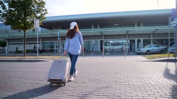 青いシャツ ジーンズ 白いキャップを着た幸せな女性のバックビューは スーツケースと空港に行く 茶色の髪の若い観光客海外で夏休みを過ごすために出発を期待 — ストック動画