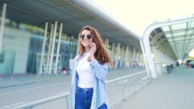 Modern binanın kentsel arka planında gezen genç ve şık yenilikçi kız öğrenci ve telefonla konuşan turist bir bavulla seyahat ediyor. Havaalanına varış ya da kalkış.