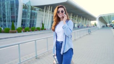 Modern binanın kentsel arka planında gezen genç ve şık yenilikçi kız öğrenci ve telefonla konuşan turist bir bavulla seyahat ediyor. Havaalanına varış ya da kalkış.
