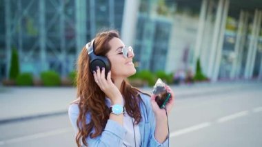 Sokaklarda müzik dinlemek için kulaklık ve modern akıllı telefon kullanan neşeli genç bir kadının portresi. Gülümseyen esmer, günlük kıyafetler ve güneş gözlüğü takıyor. Yakın plan kıvırcık hipster rahatlıyor.