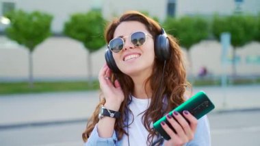 Sokaklarda müzik dinlemek için kulaklık ve modern akıllı telefon kullanan neşeli genç bir kadının portresi. Gülümseyen esmer, günlük kıyafetler ve güneş gözlüğü takıyor. Yakın plan kıvırcık hipster rahatlıyor.
