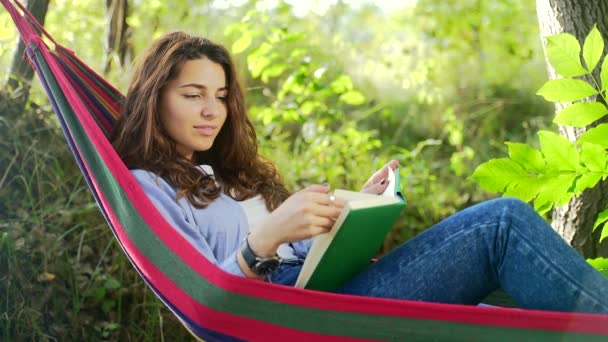 夏の公園でカラフルなハンモックに横たわって 新しい本を読んで暗い髪を持つ魅力的な若い女性 余暇と自己教育の概念 肖像画を閉じる — ストック動画