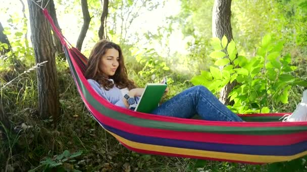 夏の公園でカラフルなハンモックに横たわって 新しい本を読んで暗い髪を持つ魅力的な若い女性 余暇と自己教育の概念 — ストック動画