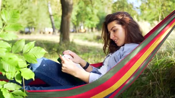 夏の公園でカラフルなハンモックに横たわって 新しい本を読んで暗い髪を持つ魅力的な若い女性 余暇と自己教育の概念 肖像画を閉じる — ストック動画