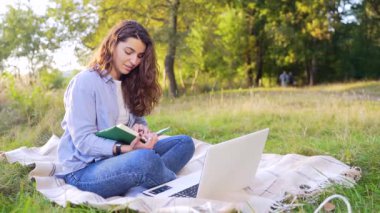 Green Park 'ta battaniyeye oturmuş modern bilgisayar eğitimi alan sıradan giysili güzel bir kadın. Koyu saçlı, açık havada deftere yazı yazan bir kız öğrenci. dizüstü bilgisayarla öğrenme 