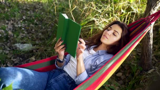 夏の公園でカラフルなハンモックに横たわって 新しい本を読んで暗い髪を持つ魅力的な若い女性 余暇と自己教育の概念 肖像画を閉じる — ストック動画