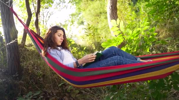 夏の公園でカラフルなハンモックに横たわって 新しい本を読んで暗い髪を持つ魅力的な若い女性 余暇と自己教育の概念 — ストック動画