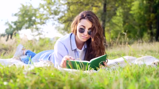 柔らかい毛布の上に横たわって 夏の公園で面白い本を読んで巻き毛の暗い髪を持つ美しい若い女性 可愛いです女性リラックス上の新鮮な空気とともに新しい文学 — ストック動画