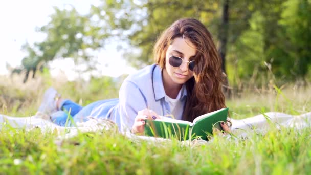 柔らかい毛布の上に横たわって 夏の公園で面白い本を読んで巻き毛の暗い髪を持つ美しい若い女性 可愛いです女性リラックス上の新鮮な空気とともに新しい文学 — ストック動画