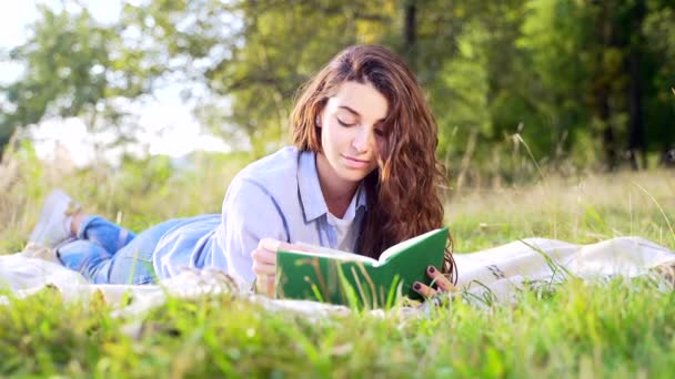 柔らかい毛布の上に横たわって 夏の公園で面白い本を読んで巻き毛の暗い髪を持つ美しい若い女性 可愛いです女性リラックス上の新鮮な空気とともに新しい文学 — ストック動画