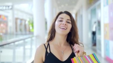 Alışveriş merkezinin önünde duran, alışveriş torbaları taşıyan ve kameraya gülümseyen neşeli genç bir kadının portresi. Kara Cuma 'da alışveriş yapan mutlu kadın neşeyle zıplar