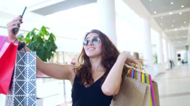 Genç, mutlu, alışveriş düşkünü bir kadının portresi elinde renkli hediye çantalarıyla akıllı telefon ya da cep telefonuyla neşe içinde selfie çekiyor. Kıvırcık, çekici bir kadın alışveriş merkezinde duruyor..