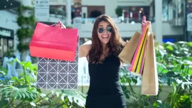 Mağazada başarılı bir alışveriş yaptıktan sonra elinde renkli poşetlerle sevinçten havalara uçan siyah saçlı hoş bir bayan. Tüketim ve alışveriş Kara Cuma mutlu genç bir kadın kameraya hediyeyle gülümsüyor.