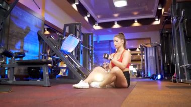 Spor kıyafetli güzel bir kadın spor salonunda sabah antrenmanından sonra dinleniyor. Yerde otururken şişeden su içen esmer bir sporcu. Güçlü bir atletik kız spor salonunda susuzluğunu giderir.