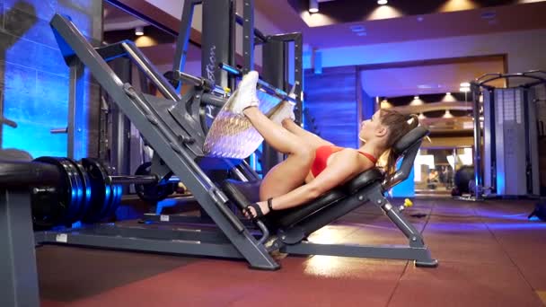 現代のジムで足のための重量機械を使用してスポーツウェアの強い若い女性 室内でフィットネストレーニングを行う茶色の髪の魅力的な女性 — ストック動画