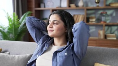 Evdeki oturma odasındaki kanepede otururken gözleri kapalı ve elleri başının arkasında rahatlayan genç ve mutlu bir kadın. Sakin, güzel bir bayan, dinleniyor, rahatlamış hissediyor. Kapat.