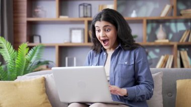Evdeki oturma odasında kanepede otururken dizüstü bilgisayardan harika haberler okuyarak başarıyı kutlayan heyecanlı genç kadın. Gülümseyen serbest çalışan kadın bilgisayardaki olumlu mesajdan memnun.