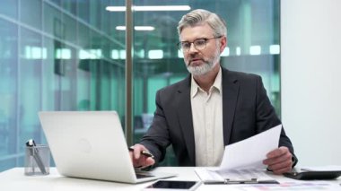 Modern iş ofisindeki çalışma masasında resmi takım elbiseli ciddi bir iş adamının portresi. Başarılı, kendine güveni tam bir finansör. Bilgisayarda çalışıyor ve kameraya bakıyor.