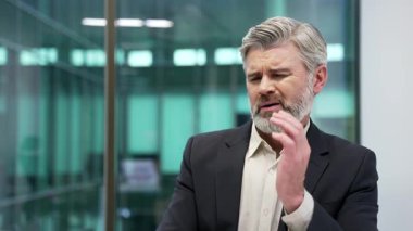 İş yerinde çalışırken diş ağrısından muzdarip olgun bir işadamı. Resmi takım elbiseli işçi elini yanağında tutuyor ve masaj yapıyor. Bir dişçiye görünmesi gerekiyor. Kapat.