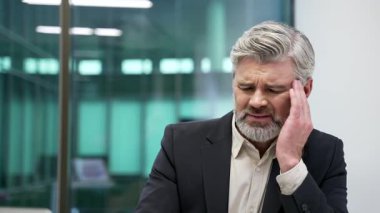 İş yerindeki iş yerinde baş ağrısı çeken olgun sakallı bir iş adamı. Resmi takım elbiseli sağlıksız işçi elleriyle şakaklarına masaj yapıyor. Çok yoruldu. Kapat.