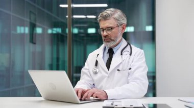Odaklanmış, beyaz önlüklü, laptopuyla hastane ofisindeki iş yerinde yazan olgun bir doktor. Düşünceli, ciddi bir tıp doktoru hastasıyla internetten bilgisayar kullanarak iş yapıyor ya da sohbet ediyor.