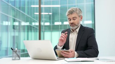 Bilgisayarla video konferansı izleyen kendine güvenen iş adamı iş ofisinde oturan not defterine notlar alır. Resmi takım elbiseli olgun sakallı yönetici online toplantıda uzaktan iletişim kuruyor.