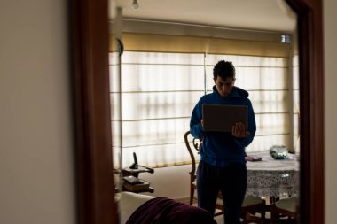 Evde bilgisayarı ve telefonuyla çalışan mavi kazaklı genç adam. 