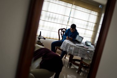 Evde bilgisayarı ve telefonuyla çalışan mavi kazaklı genç adam. 