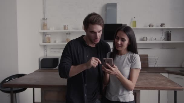 The couple in their kitchen look together at the smartphone and have fun talking, they gesticulate and smile — Stock Video